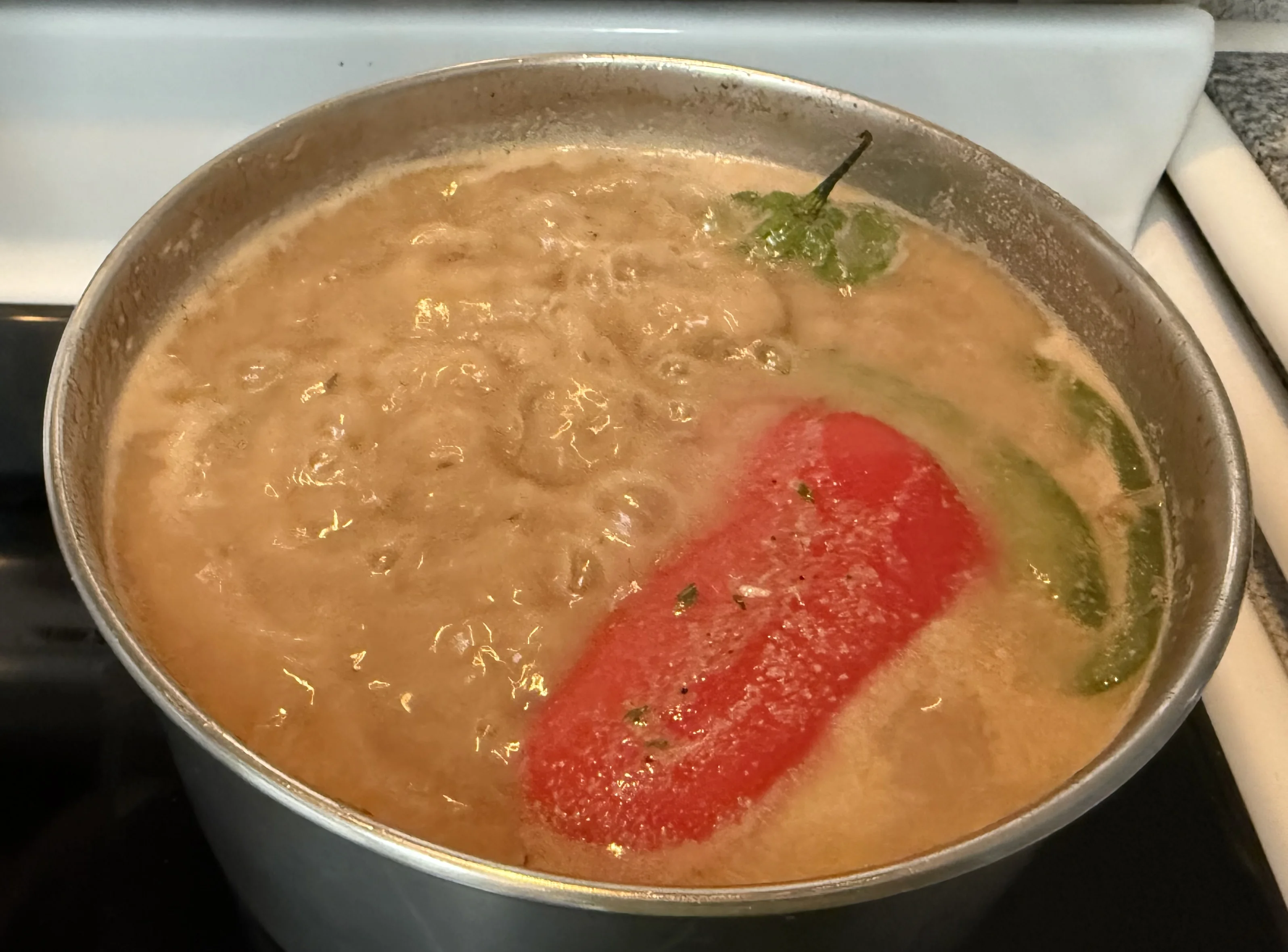 The pot simmering with coconut milk, a red pepper quarter, green pepper and a whole hot pepper