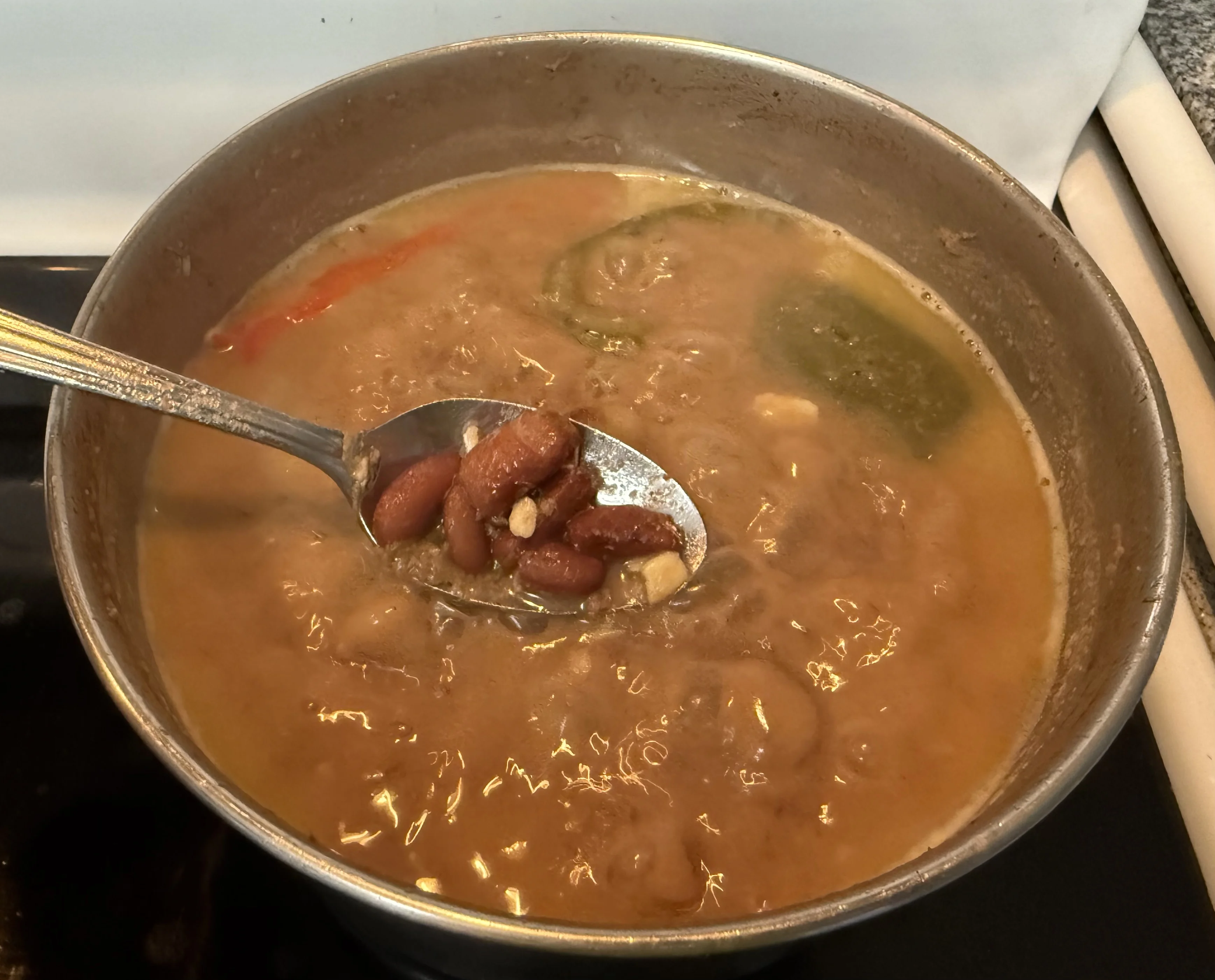 A spoon lifting cooked red kidney beans from a pot of coconut broth with peppers